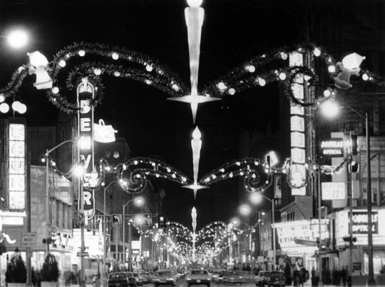 Nighttime view of 16th (Sixteenth) Street in downtown Denver, Colorado. Automobiles drive under Christmas lights and decorations that span the street. Signs read: "Denver," "Baurs," "Douglas Optical," and "Paramount."