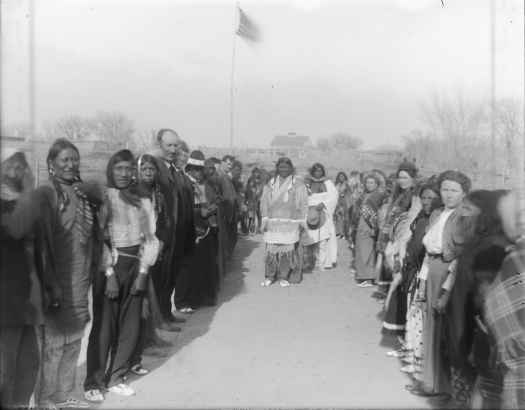 Native American (Ute) and white men and women stand in rows opposite each other probably for the traditional Bear Dance, probably in western Colorado. Shows A U.S. flag and possibly a school in the distance.