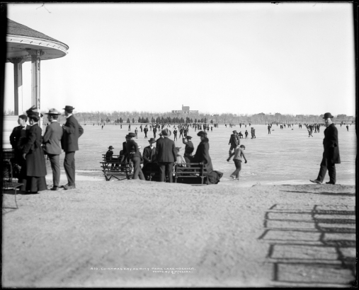 Ice skating on City Park Lake, Denver, Colorado; view east to Museum of Natural History building under construction in 1901; shows edge of City Park bandstand far left and groups seated on park bench on sandy shore.