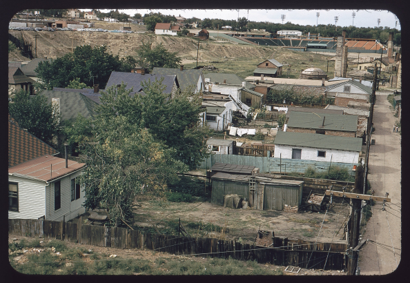 View of Bear's Stadium in the Sun Valley neighborhood, Denver, Colorado. The stadium is one level built into a hillside on the site of a landfill. Shows the backyards of houses, clothes lines, an abandoned building with smoke stacks and houses on Federal Boulevard.