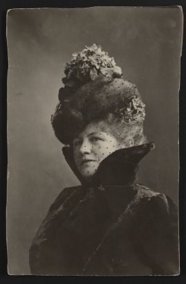 Leonel O'Bryan, otherwise known as Polly Pry, sits for a studio portrait wearing the late Victorian or Edwardian style clothing. Her outfit features a large hat adorned with silk flowers or faux foliage and a high collared coat. A sheer, black veil with chenille dots covers her face.