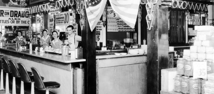 Interior view of a lunch counter at the Home Public Market, in Denver, Colorado; shows a man, women including Ruth Handler [third from right], a neon sign, tile floor, a United States flag, and signs: "Coca Cola," "Watermelon on Ice," "Beer On Draught," and "Greenwald's Soda Fountain."