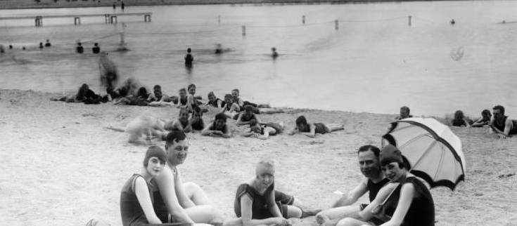 Young men and women sit on the beach at Berkeley Park in Denver, Colorado. The women wear bathing suits, bathing caps, and one woman rests a parasol over her shoulder. The men and boys nearby also wear bathing suits. Swimmers are in Berkeley Lake and on a wooden pier. The Front Range of the Rocky Mountains are is the distance.