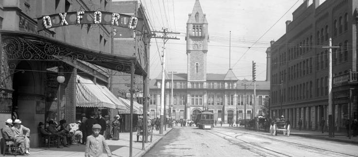 View down 17th (Seventeenth) street to Union Station prior to construction of the Welcome Arch; shows passengers boarding street railway car in center background, Oxford Hotel at left, Hendrie and Bolthoff Manufacturing and Supply Company at right, horse-drawn carriages on the street, and a young boy with knickers approaching bicycle left foreground; many men sit in front of the Oxford Hotel.
