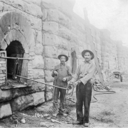 Two workers stand near a row of coke ovens in the coal camp of El Moro (Las Animas County), Colorado. A man wearing overalls holds a rake. The other man holds a pole that extends into the opening of an oven. The men may be Italian or Hispanic. Wheelbarrows, ladders, and other tools are in the distance.