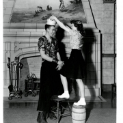 Photograph of Adolph Mitterer, as he is crowned the winner of a "Beauty Contest" held at West High School in Denver, Colorado.  Juanita Burke, another student at the school puts a paper crown on his head.  Burke is standing on a small wooden barrel and step stool to place the crown.  Mitterer is wearing a floral blouse, long black skirt and army boots.