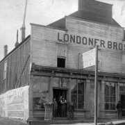 Men stand and pose near doorways at the wood frame Londoner Bros. store with a gabled roof and a false front in Leadville (Lake County), Colorado. Men wear hats, suits or vests, and stand beside store windows and a straw broom display on the wooden sidewalk. A painted sign reads "Londoner Bros. Groceries & Mining Supplies." A poster on the side of the building reads: "Barry & Fay in their new comedy, Irish Aristocracy." A woman wears a dress, a hat and holds a parasol on the street near the store.