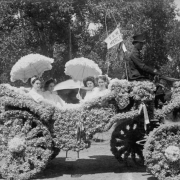 A Pumpkin Pie Day Parade float pauses on a street, Longmont, Colorado. Hattie Secor, Martha Donova, Louise Boynton and Harriet Smith sit in the carriage elaborately decorated with flowers. They wear white dresses and hold parasols with ruffles. Rudy Garner, the driver in a top hat, holds the reins of the horse.