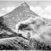 View of a smoking locomotive with passenger cars on the standard- gauge Colorado Springs & Cripple Creek District Railway; St. Peter's Dome rock formation in background with flag flying.