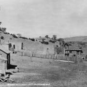 View of adobe houses in a Mexican settlement near Walsenburg (Huerfano County), Colorado. Shows a man by a horse, beehive ovens (hornos), a wooden fence, a wagon with a barrel on it and a cedar tree.