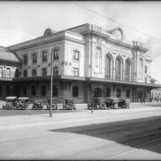 Northwestward, exterior view of the Union Station train depot in Denver, Colorado; shows automobiles parked in front of station, a boy standing on sidewalk, streetcar tracks on Wynkoop Street, and people standing in front of station at far right.