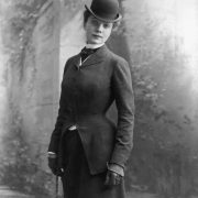 Standing studio portrait of Alma Regina Shelgram Osgood, second wife of John Cleveland Osgood, mining magnate of Redstone, Colorado. She is fashionably dresses in a fitted ridding jacket, bowler hat and gloves. A painted garden wall scene serves as the backdrop.