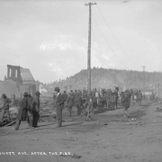 Large group of men and on-lookers view destruction, debris and remains of two-story brick wall on Bennett Avenue caused by second fire on April 29, 1896, Cripple Creek, Colorado; mule-drawn wagon loaded with wooden barrels is parked along edge of street; men are lined along railing by the elevated portion of Bennett Avenue; Tutt-Penrose building and other commercial business buildings untouched by fire.