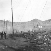 Destruction west on Bennett Street caused by second fire on April 29, 1986, Cripple Creek, Colorado; groups of men and two men on horseback in street survey rubble and remains of once thriving commerical business district; shows utility poles with fallen wires, chimneystack, remains of two-story brick building, Mt. Pisgah is on horizon and untouched  residences are on hillside in background.