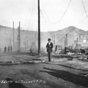 View of destruction west on Bennett Avenue, Cripple Creek, Colorado, caused by second fire on April 29, 1896; groups of men and solitary men survey remaining debris; smoke still rises from ashes and utility poles with fallen wires border street; Mt. Pisgah and undamaged buildings are in background.