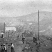 Aftermath of destruction caused by April, 1896 fire, Cripple Creek, Colorado; men, women and children stroll down street that has debris  and rubble left from fire; foreground includes woman holding baby, boy drinking from fire hydrant, canvas tents amidst rubble, and stream of water  sprayed on rubble.