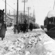 Pedestrians, a man carrying skis, horse drawn wagons and street cars number 92 and 75 make their way through deep snow on Colfax Avenue, Denver, Colorado. The Argonaut Hotel and the Immaculate Conception Cathedral show in distance.