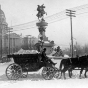 A man hauls his wagon load of snow past the Pioneer Monument on Cheyenne Street towards Civic Center, Denver, Colorado. The Scholtz Drug Company, Capitol Store, the State Capitol Building and the First United Presbyterian Church show in distance.
