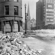 View from corner in front of the Brown Palace north along Broadway, Denver, Colorado, includes piles of snow on streets, the Hotel Metropole and the Trinity Church. Sign painted on side of hotel reads: "Hotel Metropole American and European Plan, Absolutely Fire Proof."