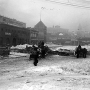 Pedestrians, horse drawn wagons and sleighs make their way through a snow filled Cheyenne Street, Denver, Colorado. The Y. M. C. A. and Plymouth Hotel show in background. Signs read: "Fry and McHill," "Baker Electrics?" "Ford Motor Company," and "The Wilson Auto Co."