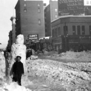 A young boy poses in front of a tall snowman next to a snowcovered post, downtown Denver, Colorado. Snow piles have American flags stuck in them for decoration. The sidewalks are cleared, while the street shows deep snow heavily tracked. Signs read: "Clarke's Restaurant" and "Tom Moore, Cigar 10 cents, Little Tom 5 cents."