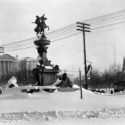 View of deep snow from Cheyenne Street towards Pioneer Monument and the State Capitol Building, Denver, Colorado. A workman repairs wires on a power and streetlight pole. The Scholtz Drug Company is on the corner; trolley cars and the United Presbyterian Church shows in background.