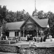 Tourists come and go from the Soda Spring Pavilion, Manitou Springs, El Paso County, Colorado. The open air pavilion features decorative woodwork with sunburts, rusticated stone foundation, roof cresting, four sided dome with flag pole and fish scale shingles. Signs read: "Manitou Springs," and "Developing and Finishing Films and Supplies."