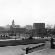 View looking northwest towards downtown from State Capitol steps, Denver, Colorado, includes cable street cars, carriages, Engine Company No. 1's victorian firehouse across the intersection of Broadway and Colfax and the Arapahoe County Courthouse in distant haze. Signs on billboards and foreground businesses read: "Palace Stables," "Plymouth Hotel," "$25.00 Rio Grande," "Denver Clothes Pressing Co., $3.00 for 3 Months," "Automobiles, Repairs, Storage."