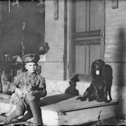 Outdoor portrait of a boy in Denver, Colorado, wearing a World War One or Western Union Telegraph uniform. He holds a tabby cat; an Irish Setter is by a brick wall with graffiti.