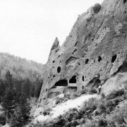 A Native American man stands next to a tuff cliffside with eroded holes enlarged by ancestral Pueblo Indians for dwellings and ceremonial spaces, Pajarito Plateau, New Mexico; became Bandelier National Monument in 1916.