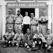 The Pine Crest Baseball team poses in front of the Palmer Lake Bowling Alleys, El Paso County, Colorado. They wear uniforms (caps, shoes with cleats, baseball gloves.) The catcher with his mitt sits front and center; players next to him have wooden bats. Possibly the coach or owner of the bowling alley stands in center back row. He wears a white shirt and striped tie.