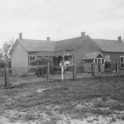 People pose on the porch of Senator Casimiro Barela's house in probably Barela (Las Animas County), Colorado.  Shows a cross gabled adobe house with a covered porch, wood and wire fence and out buildings.