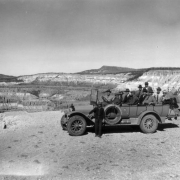 Group shot of possibly President Harding and cabinet members on their 1923 tour of Yellowstone National Park, Wyoming. The driver stands beside the long touring automobile filled with men, parked on at a lookout over eroded canyon cliffs. Emblem on door has a bear and reads: Y. P. T. Co. No. 143.
