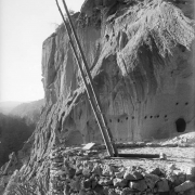 A timber ladder provides access into a kiva structure under volcanic tuff cliffs, Frijoles Canyon, Bandelier National Monument, New Mexico. The reconstructed kiva is built up with rock and has a rectangular entrance framed with timber. A natural cave enlarged by ancestral Pueblo Indians shows in background.