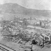 Rubble and destruction caused by second fire of April 29, 1896, Cripple Creek, Colorado; piles of twisted sheet metal, canvas tents amidst rubble, railroad tracks in foreground; view across fire devastated area toward distant view of remaining buildings; horse-drawn wagons and carriages on street.