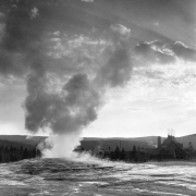 View southwest over Old Faithful Geyser to Old Faithful Inn, with steep gabled roof and a large rooftop observation platform with six flagpoles, Upper Geyser Basin, Yellowstone National Park, Wyoming. Tourists climb the terraces around the steaming geyser.