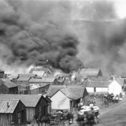View towards El Paso Livery Stable, Bennett Avenue between 1st (First) and "A" Streets, consumed in flames and smoke after explosion of stored dynamite at stable during second fire on April 29, 1896, Cripple Creek, Colorado; billows of black smoke cover view of town and numerous horse-drawn wagons packed with personal belongings are parked in front of wood frame buildings and residences; scence shows blurred images of moving wagons and unhitched carriages.