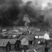 View towards El Paso Livery Stable, Bennett Avenue between 1st (First) & "A" Streets, as fire causes stored dynamite to explode during second fire on April 29, 1896, Cripple Creek, Colorado; actual blast pictured with roof debris in air, black smoke covering view of town, numerous wood frame residences and buildings in path of fire, and horse- drawn wagons packed with personal belongings; families watch approaching smoke and flames.