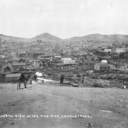 View west from Myers Avenue (probably) towards Bennett Avenue with Mt. Pisgah on horizon after first fire of April 25, 1896, Cripple Creek, Colorado; scene includes debris and  remains from fire, construction  of wood frame buildings and canvas tents and a distant view of numerous remaining wood frame and brick commercial buildings in business district; signs on buildings include: "Metropolitan House," "El Paso Livery," "Drugs," and "One price to All-King Cash-Our Motto-Weinberg Shoes & Clothing Co."