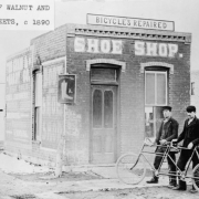 Men hold a bicycle-built-for-two in front of a brick commercial building in Fort Collins, Larimer County, Colorado. Signs read: "Bicycles Repaired," and "Shoe Shop;" another depicts a boot and reads: "Ludincton & Co."