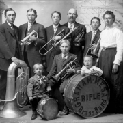 Members of the Graham (family) Mesa Brass Band pose with musical instruments in Rifle (Garfield County), Colorado. They are identified as (top row, left to right): Elmer, Albert, Claude, Isem W., Henry and Jennie and (bottom row, left to right): Vern, Eber, and Wilbur. The members of the family band hold musical instruments which include trumpets, trombones, tubas, and drums. The man and the boys wear suits and ties. Letters on a bass drum read: "Rifle Graham Mesa Brass Band."