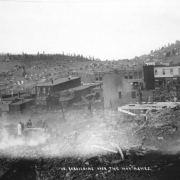 View of smouldering remains from first fire on April 25th, 1896, Cripple Creek, Colorado; three men look over debris and one man sprays water over building remains; flat roof brick and wooden frame false front buildings and the Tutt & Penrose building are in the commercial district on  Bennett Avenue; wood frame residences are on hillside in the background.