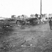 Destruction on First Street caused by second fire, April 29, 1896, Cripple Creek, Colorado; remains include stone or cement block foundations, clustered groups of high-backed chairs, tables, stool, wood commode and wagon wheels; man walks down dirt street; one-story gabled and hipped roof residences are in background.