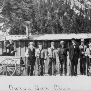 The Ouray Gun Club poses with their rifles outside a board-and- batten shack on a snowy day north of Ouray, Ouray County, Colorado. They include, left to right: Gus Arps in cart drawn by a small burro, unidentified man, Albert Arps, Otto Arps, Charley Pearson, Barney Du Praw, Andy Sneva, Ed Arps, Alf Armstrong, George Armstrong (little boy.)