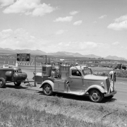 View of the Remington Ordnance Plant (later called the Federal Center) in Lakewood (Jefferson County) Colorado; men install road reflectors by a (probably) striping truck, with a compressor in tow. Sign reads: "War Department - Quartermaster Corps - Construction Division - Denver Ordnance Plant - Smith Henchman & Grylls, Inc., Broderick and Gordon General Contractors." Front range mountains and clouds are in the distance.