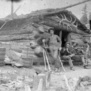 Three men pose by a log cabin belonging to S. J. Roberts, Cripple Creek, Colorado; one man with pants tucked into knee-high boots stands near miner's pick, broom, and sledge hammer (possibly S. J. Roberts); men sit holding a flute or fife and a fiddle or violin; a rack of elk's antlers is over the door.