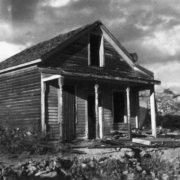 The Simmons house, on Letcher Street, in Nevadaville, Gilpin County, Colorado, is a dilapidated one story frame structure with a gable roof, a covered porch, and lathe-turned piers. Door and window openings are  empty or covered by corrugated tin; debris covers the ground, and ruins of  stone walls are in the background.
