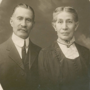 Studio portrait of Colorado State Senator from Las Animas County, Colorado, Casimiro Barela and his wife Damiana Rivera de Barela. Senator Barela wears a suit with an oversized celluloid collar and necktie. He has a trimmed, gray mustache and his graying hair is combed straight back. His wife, Damiana, wears a dress with a light colored insert and a high, lace collar. She has on diamond earrings and a pin with a pendant and her gray hair is combed back and parted in the middle.
