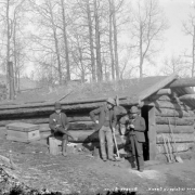 First house in Poverty Gulch, Cripple Creek, Colorado; house is possibly Robert Womack's cabin in Fremont (prior to Cripple Creek incorporation) or Andy Frazier's cabin in Squaw Gulch; the one-story rounded log cabin built into the hillside has a low gable sod roof;  three men wearing three-piece suits and knee high boots pose outside cabin; one man leans on miner's pick while another man, wearing a derby hat, sits on stump; additional picks and shovel are near front side entrance of cabin.
