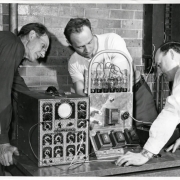 Photograph of three men during a radio repair class at the Emily Griffith Opportunity School located in Denver, Colorado.  Two men watch while a man in the center stands behind the machine working on it.